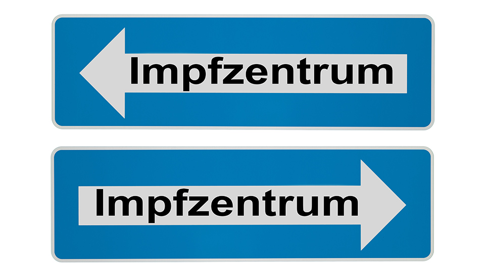 Zwei Verkehrsschilder in Blau, auf dem jweils ein weißer Pfeil mit der Aufschrift "Impfzentrum" steht. Die beiden Pfeile zeigen in eine unterschiedliche Richtung