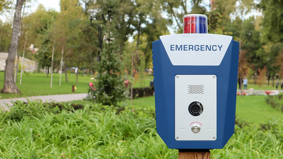 Blau-weiße Emergency-Notruf-Säule im Vordergrund. Im Hintergrund ein Park mit viel Grünfläche.