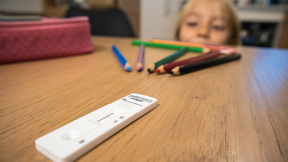 Karierter Block im Hintergrund und im Vordergrund eine Kinderhand, die einen Corona-Test hält. Unter diesem Test liegt ein bemaltes Papierherz.