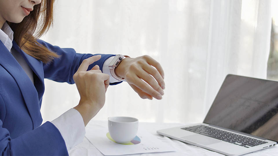 Ausschnitt einer Frau, die gerade mit dem Finger auf ihre Smartwatch tippt. Im Hintergrund steht ein aufgeklappter Laptop und eine weiße Tasse.