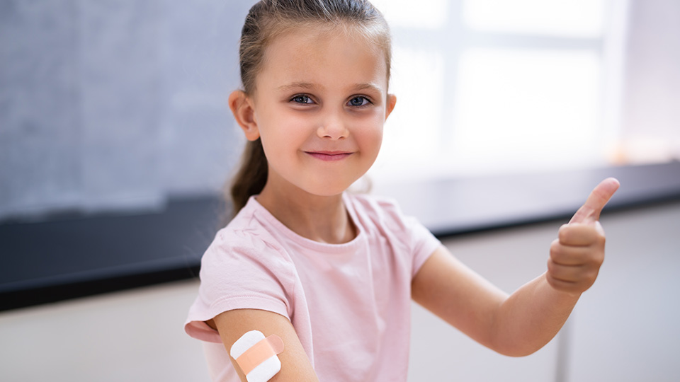 Mädchen mit rosa Oberteil und einem Pflaster am Arm, macht das Daumen-Hoch-Zeichen nach ihrer Covid 19 Impfung.