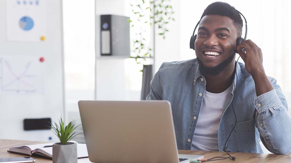 Junger Mann, der mit einem Headset vor seinem Laptop sitzt und lacht.