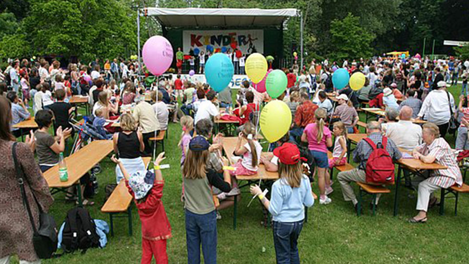 Kinderbürgerfest: Erwachsene und Kinder mit Luftballons im Stadtpark, dahinter eine Bühne