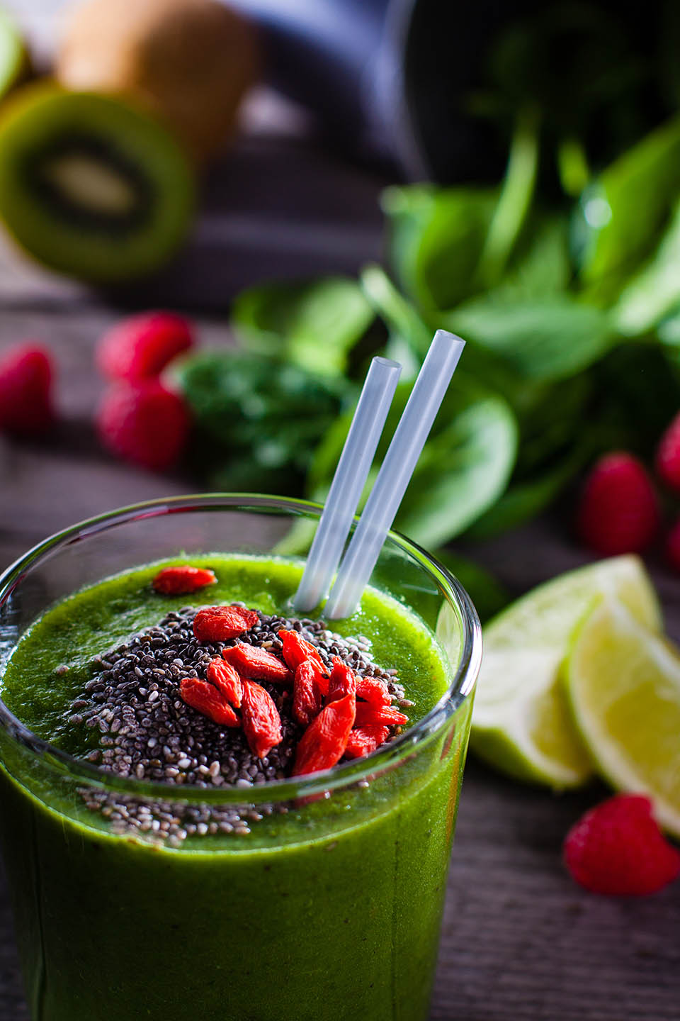 Fresh green smoothie sprinkled with chia seeds and goji berries in the background fruits and vegetables on wooden table.