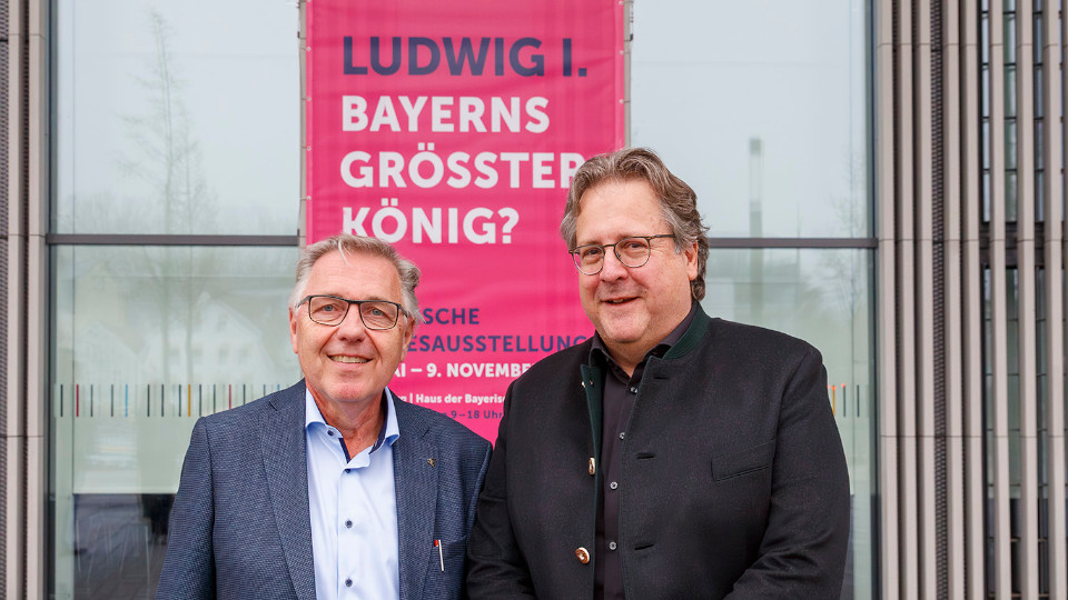 Dr. Rainhard Rieperting und Dr. Richard Loibl mit dem enthüllten Fassadenbanner zur Bayerischen Landesausstellung 2025 „Ludwig I. – Bayerns größter König?“.